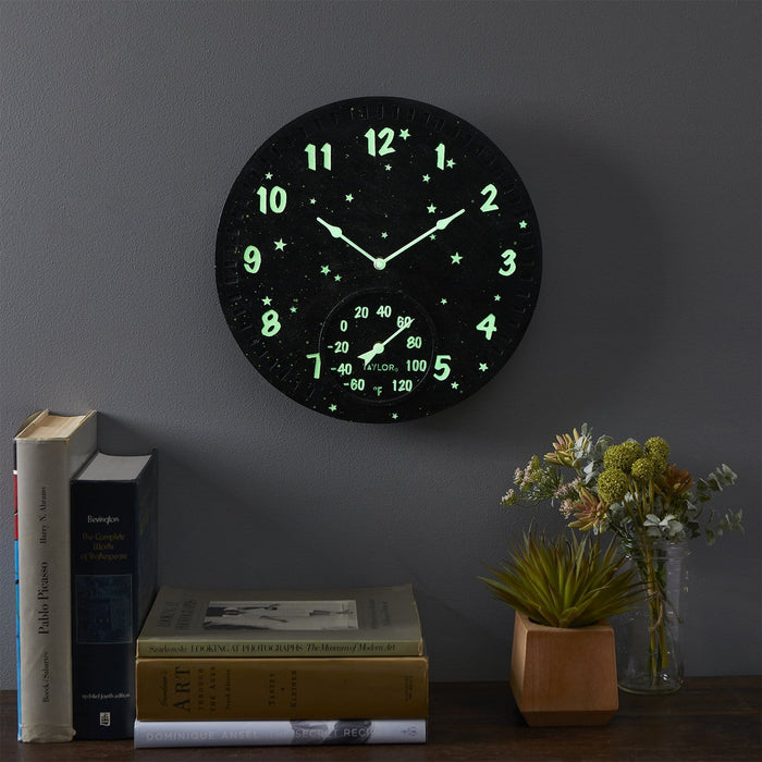 Wall mounted 14 inch Galaxy Glow in the Dark Clock with Thermometer glowing on a dark wall beside books and plants