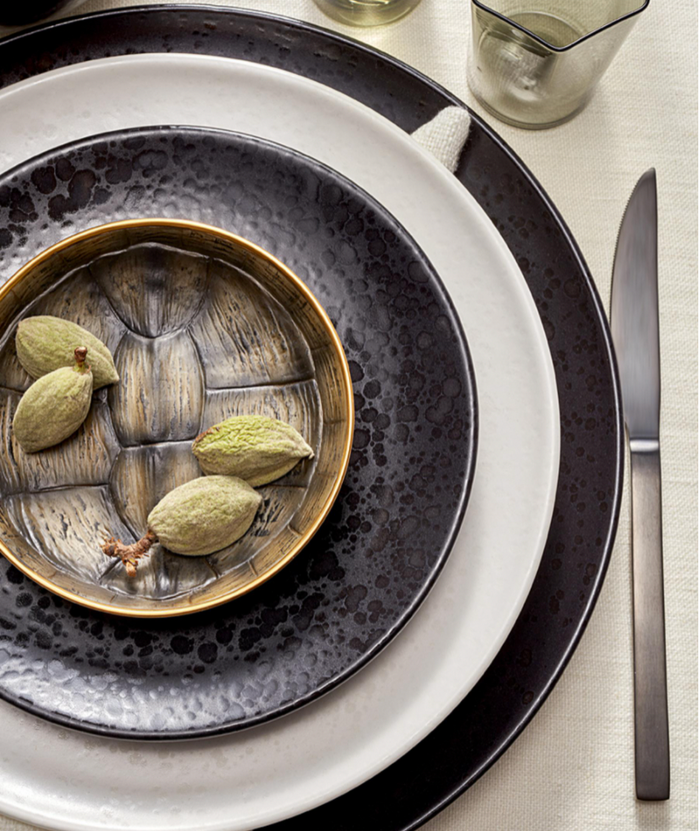 A rustic place setting featuring three L'Objet Alchimie Dinnerware White plates, a small metallic bowl filled with green almonds, and a dark gray knife.