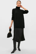 Woman in a classic Black Louisa Turtleneck Sweater with ribbed trims, paired with a satin midi skirt, knee-high boots, and gold accessories for an effortless, high-end look.