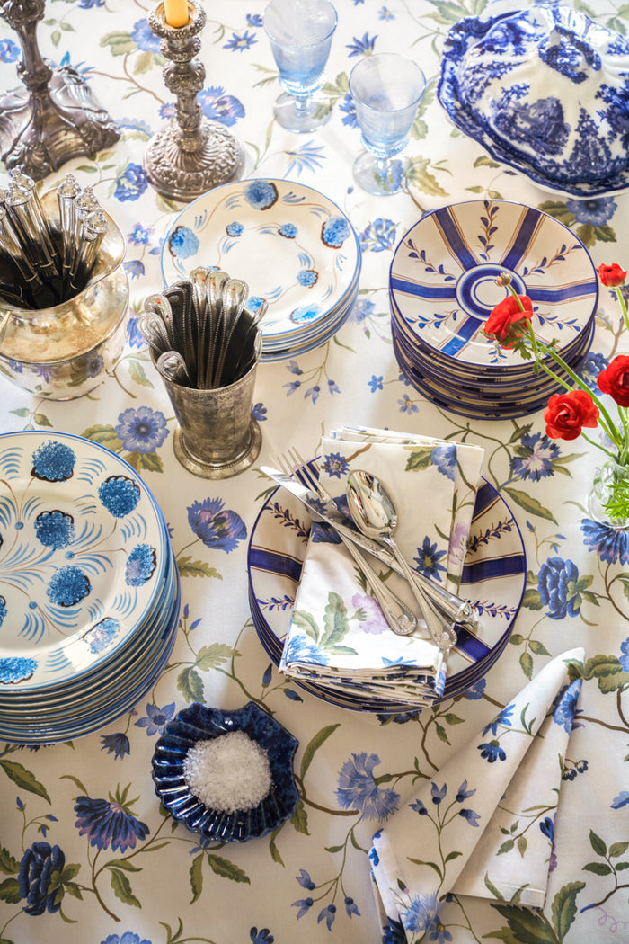 Elegant tabletop featuring the Blue Flower Dinner Plate, hand-painted in Portugal, set amidst classic floral linens, vintage silver, and vibrant decor for an effortlessly high-end dining experience.