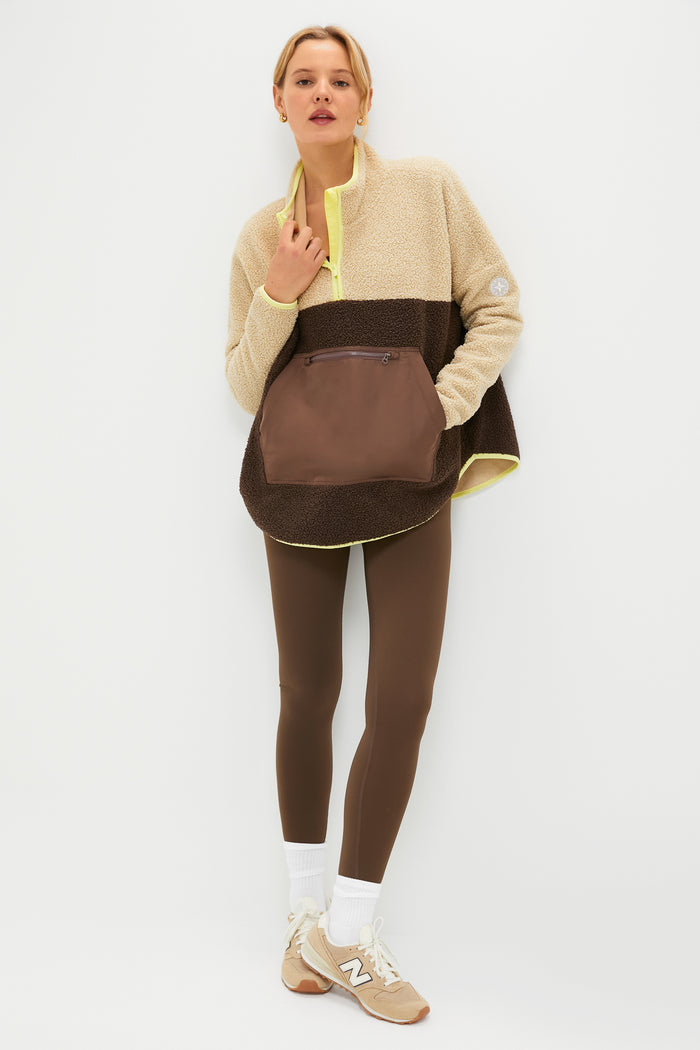A woman with short hair stands against a white wall, wearing a Tnuck Sport beige and brown fleece pullover with a kangaroo pocket, brown leggings, white socks, and beige sneakers. Her hands are tucked into the pullover’s pocket.