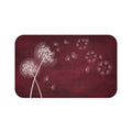 This is an image of a rectangular floor mat with rounded corners. The background of the mat mimics distressed, burgundy chalkboard. The primary graphic is a cluster of stylized dandelion seed heads on the left side. The seed heads are drawn in fine detail, and several dandelion seeds are shown floating across, suggesting a gentle breeze.