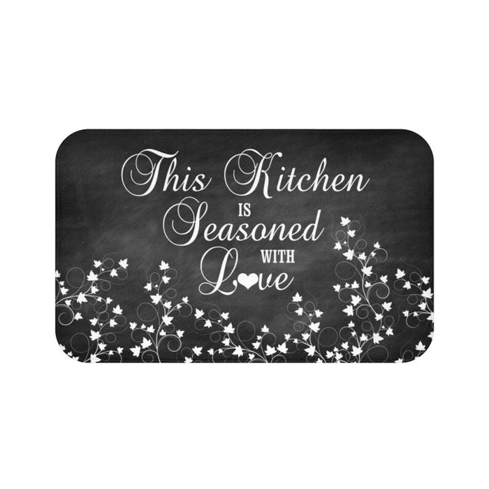 A black chalkboard motif background on a rectangular kitchen memory foam mat with the text 'This Kitchen is Seasoned with Love' and vine scroll graphics across the bottom half of the mat in a white cursive font. The "o" in the word "Love" is replaced with a heart  symbol.