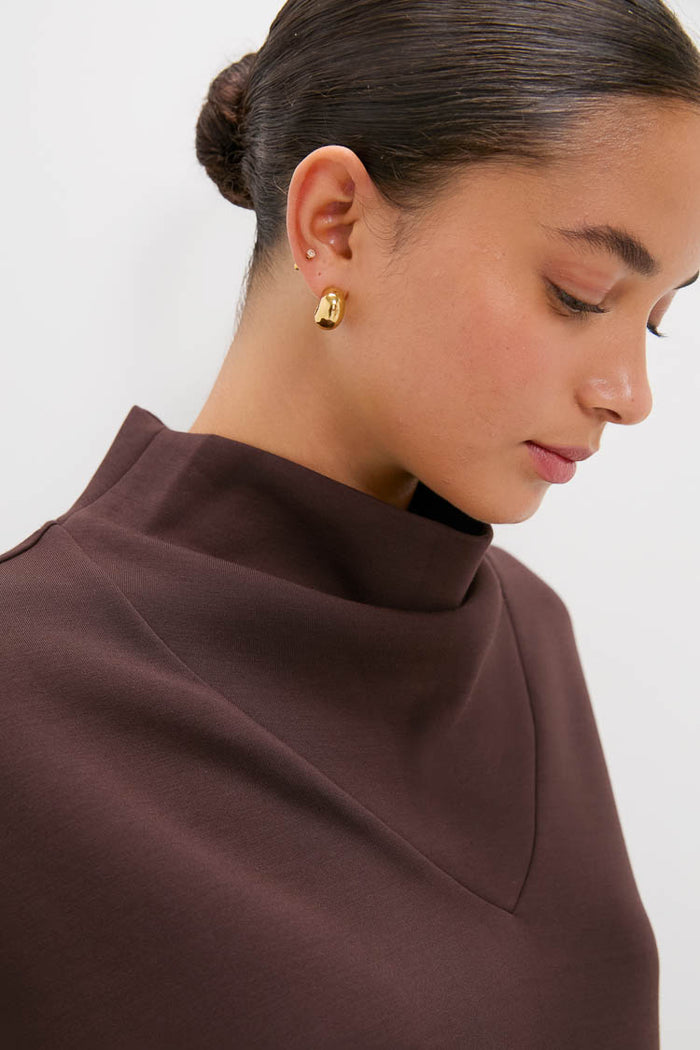 A woman with dark hair in a bun wears gold oval earrings and a brown Varley top featuring a structured high neckline. She is facing slightly downward against a plain background.
