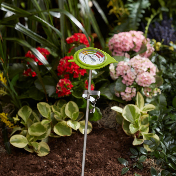 Close up of a lime green compost thermometer with decorative leaf guard in a garden with red and pink flowers in the background.