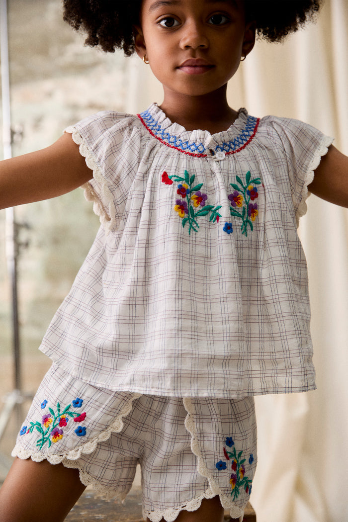 Young child wearing the classic Cream Cruise Checks Perlane Blouse with smocked neckline and floral embroidery, paired with matching shorts. Effortlessly fun and high-end style.