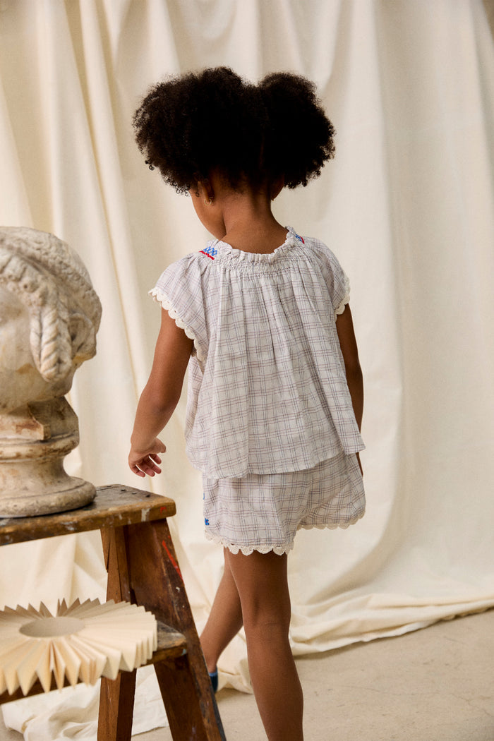 Child in classic Cream Cruise Checks Perlane Blouse and shorts, featuring embroidered details, stands by a wooden stepladder in a softly lit studio, embodying effortless, high-end boho style.