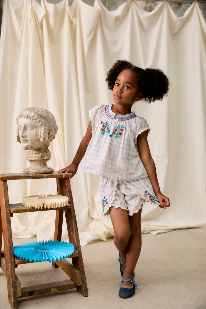 Preschool girl in Cream Cruise Checks Perlane Blouse with scalloped shorts, standing beside stool with classical bust and paper fans. Effortless, classic, and high-end boho style.