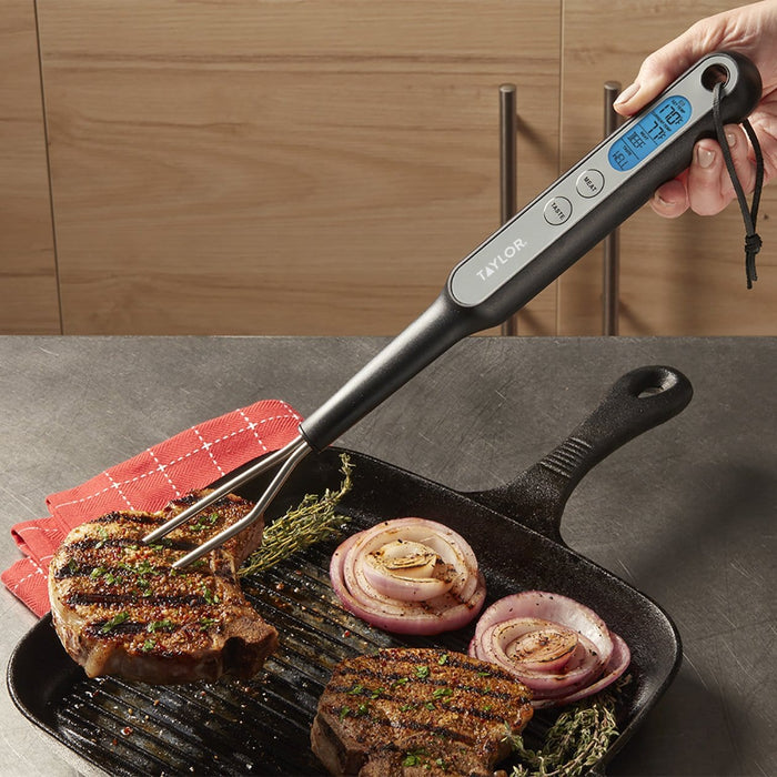 Taylor digital fork thermometer with blue backlit display held over a skillet grilled steak and onions.