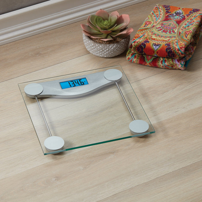 Digital glass scale with clear metal accents on a light wood floor beside a potted succulent and a colorful blanket.