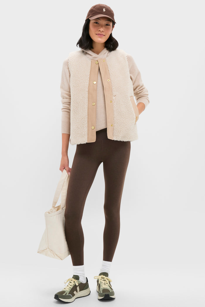A woman wears a brown cap, Barbour Dulsie Liner beige fleece vest, light hoodie, brown leggings, white socks, and sneakers, holding a cream tote bag—showcasing a cozy Barbour look against a plain background.