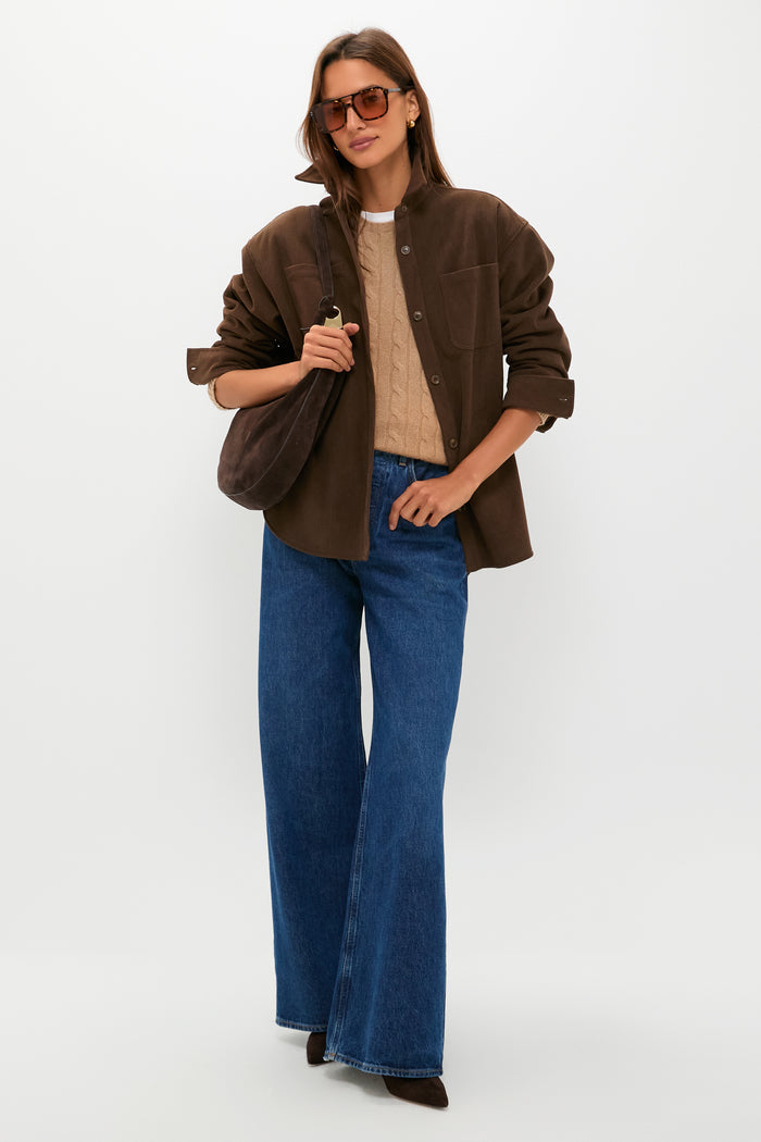 A woman in Veronica Beard oversized sunglasses, a brown suede jacket, a beige sweater, blue wide-leg jeans, and brown pointed kitten heels stands against a plain white background.