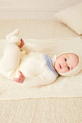 A baby rests on a cream blanket, wearing a cozy Pepa London Fair Isle baby set made from soft merino wool with blue details and a matching hat, on a light carpeted floor.
