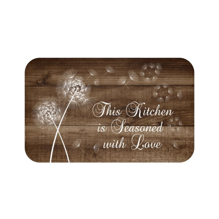 Memory foam floor mat with a brown wood plank background and a white dandelion pattern with the dandelions drifting in the wind to the right side of the mat. The phrase "This Kitchen is Seasoned with Love" is in the lower-right section of the mat.