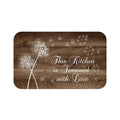 Memory foam floor mat with a brown wood plank background and a white dandelion pattern with the dandelions drifting in the wind to the right side of the mat. The phrase "This Kitchen is Seasoned with Love" is in the lower-right section of the mat.