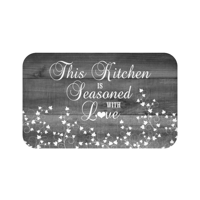 A gray wood plank background on a rectangular kitchen memory foam mat with the text 'This Kitchen is Seasoned with Love' and vine scroll graphics across the bottom half of the mat. Text and vine scroll are white. The "o" in the word "love" is replaced with a heart shape.