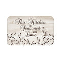 A sand-colored wood plank background on a rectangular kitchen memory foam mat with the text 'This Kitchen is Seasoned with Love' and vine scroll graphics across the bottom half of the mat. Text and vine scroll use a dark brown color.