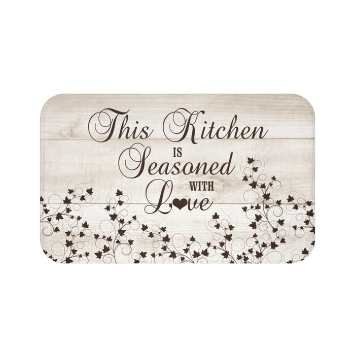 A sand-colored wood plank background on a rectangular kitchen memory foam mat with the text 'This Kitchen is Seasoned with Love' and vine scroll graphics across the bottom half of the mat. Text and vine scroll use a dark brown color.