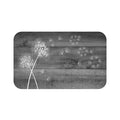 This is an image of a rectangular floor mat with rounded corners. The background of the mat mimics distressed, gray wooden planks. The horizontal plank lines give the mat a texture and farmhouse feel. The primary graphic is a cluster of stylized dandelion seed heads (in white) on the left side. The seed heads are drawn in white, fine detail, and several dandelion seeds are shown floating across, suggesting a gentle breeze.