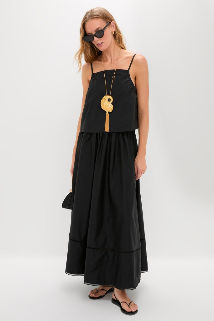 Woman in an effortless, high-end ensemble wearing the Gold Aveline Necklace, a fan-shaped pendant with tassel, paired with a classic black outfit and small handbag.