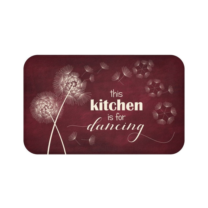 This is an image of a rectangular floor mat with rounded corners. The background of the mat mimics distressed, burgundy chalkboard. The primary graphic is a cluster of stylized dandelion seed heads on the left side. The seed heads are white and drawn in fine detail, and several dandelion seeds are shown floating across, suggesting a gentle breeze. The design is enhanced with the phrase "this kitchen is for dancing" in mixed fonts.