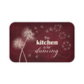 This is an image of a rectangular floor mat with rounded corners. The background of the mat mimics distressed, burgundy chalkboard. The primary graphic is a cluster of stylized dandelion seed heads on the left side. The seed heads are white and drawn in fine detail, and several dandelion seeds are shown floating across, suggesting a gentle breeze. The design is enhanced with the phrase "this kitchen is for dancing" in mixed fonts.