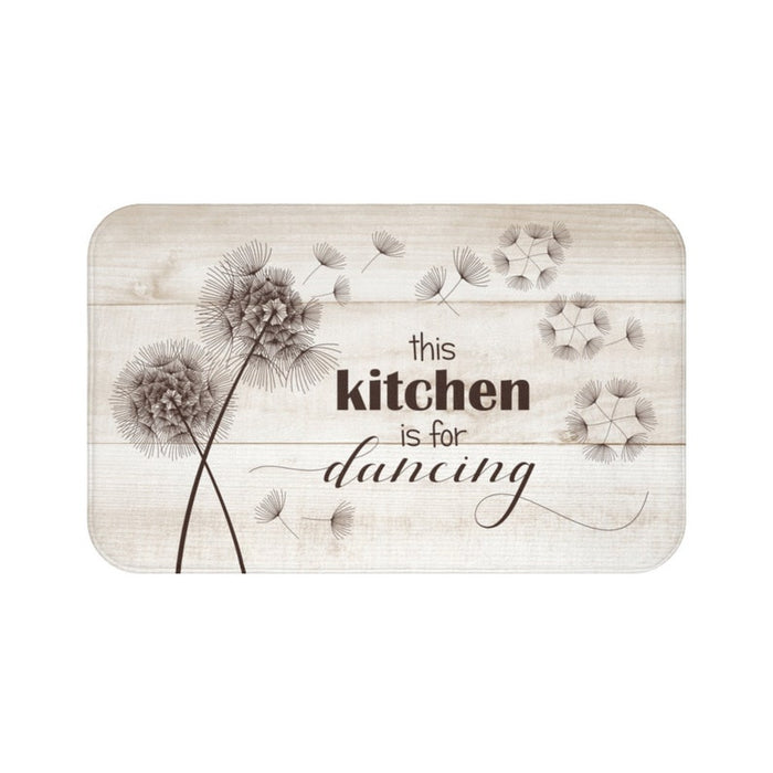 This is an image of a rectangular floor mat with rounded corners. The background of the mat mimics distressed, tan-colored wooden planks. The horizontal plank lines give the mat a texture and farmhouse feel. The primary graphic is a cluster of stylized dandelion seed heads on the left side. The seed heads are drawn in white, fine detail, and several dandelion seeds are shown floating across, suggesting a gentle breeze. The design is enhanced with the phrase "this kitchen is for dancing" in mixed fonts.