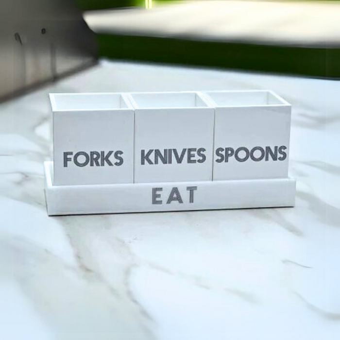 The Eye Candy Let's Eat Silverware Caddy in white with dark grey has three labeled sections for forks, knives, and spoons, and the word "EAT" on the base. It sits stylishly on a marble countertop.