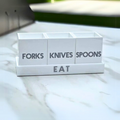 The Eye Candy Let's Eat Silverware Caddy in white with dark grey has three labeled sections for forks, knives, and spoons, and the word "EAT" on the base. It sits stylishly on a marble countertop.