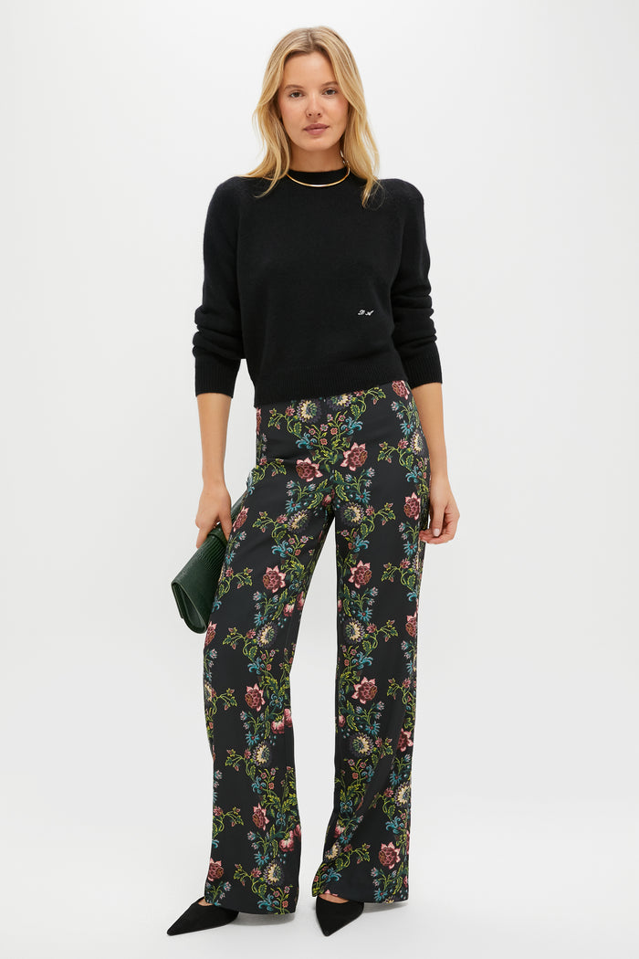 A woman stands against a white background wearing a Tuckernuck black sweater, wide-leg floral trousers, black pointed shoes, and holding a small green clutch.