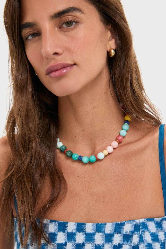 A woman models the Multi Bright By Nature Necklace, a classic and fun chunky, multicolored beaded piece, paired effortlessly with a gold hoop earring and checked tank top.