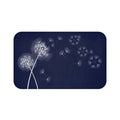 This is an image of a rectangular floor mat with rounded corners. The background is solid navy/deep blue. The primary graphic is a cluster of stylized dandelion seed heads on the left side. The seed heads are drawn in white, fine detail, and several dandelion seeds are shown floating across, suggesting a gentle breeze.