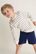 Young boy smiling in a classic Navy Stripe Raya Cotton Jumper, featuring ribbed trims and wooden shoulder buttons. Effortless and fun, perfect for high-end style.