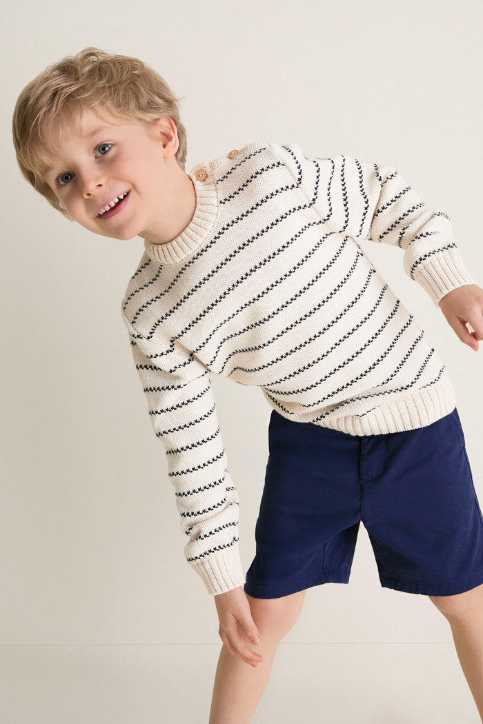 Young boy smiling in a classic Navy Stripe Raya Cotton Jumper, featuring ribbed trims and wooden shoulder buttons. Effortless and fun, perfect for high-end style.