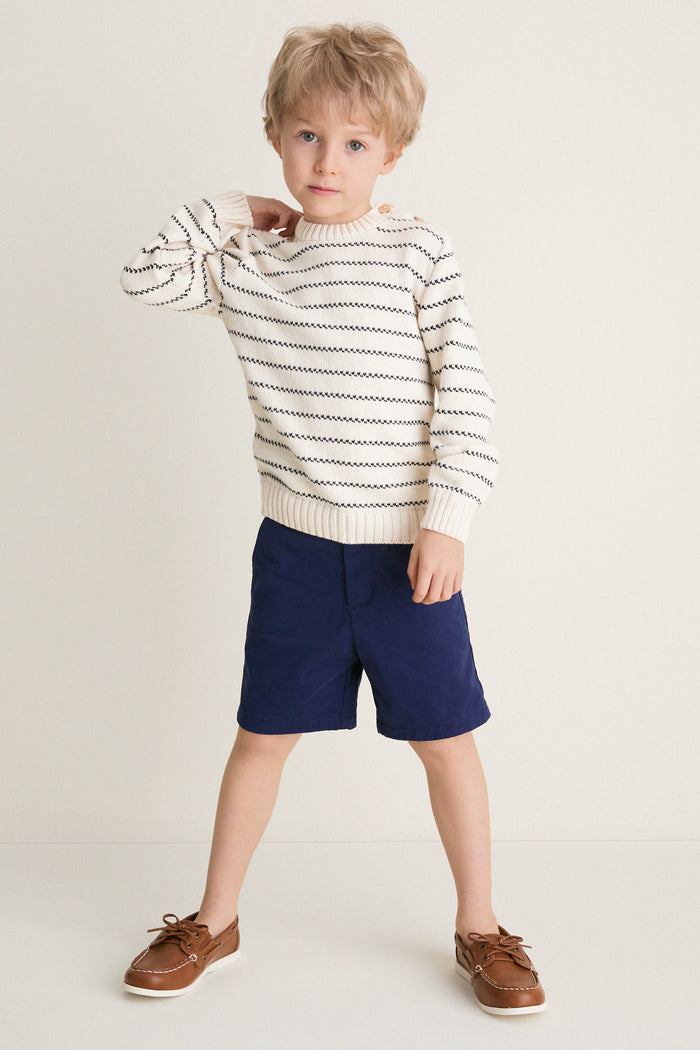 Young boy in a classic Navy Stripe Raya Cotton Jumper, showcasing effortless style with ribbed trims and wooden buttons, paired with navy shorts and brown boat shoes.