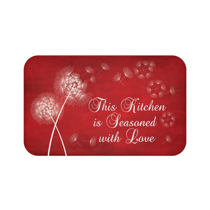 A red rectangular kitchen memory foam mat with white text and dandelion designs, and the phrase 'This Kitchen is Seasoned with Love'.