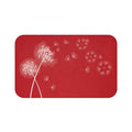 This is an image of a rectangular floor mat with rounded corners. The background is solid red. The primary graphic is a cluster of stylized dandelion seed heads on the left side. The seed heads are drawn in white, fine detail, and several dandelion seeds are shown floating across, suggesting a gentle breeze.