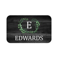 A rectangular black memory foam mat with a green wreath and single letter monogram design in the center, and the personalized name 'Edwards' at the bottom as an example of the personalization options.
