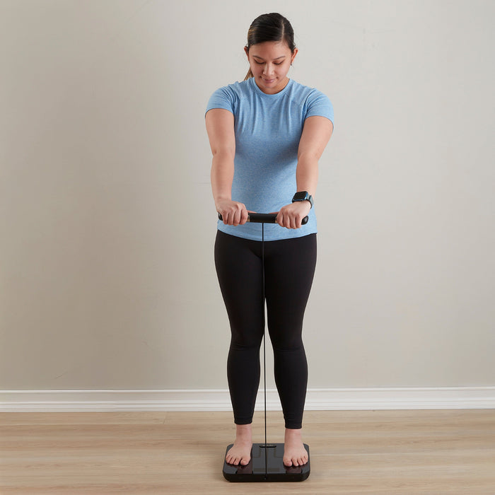 Person wearing a blue athletic top stands on a smart total body composition scale on a wooden floor, wearing a smartwatch.