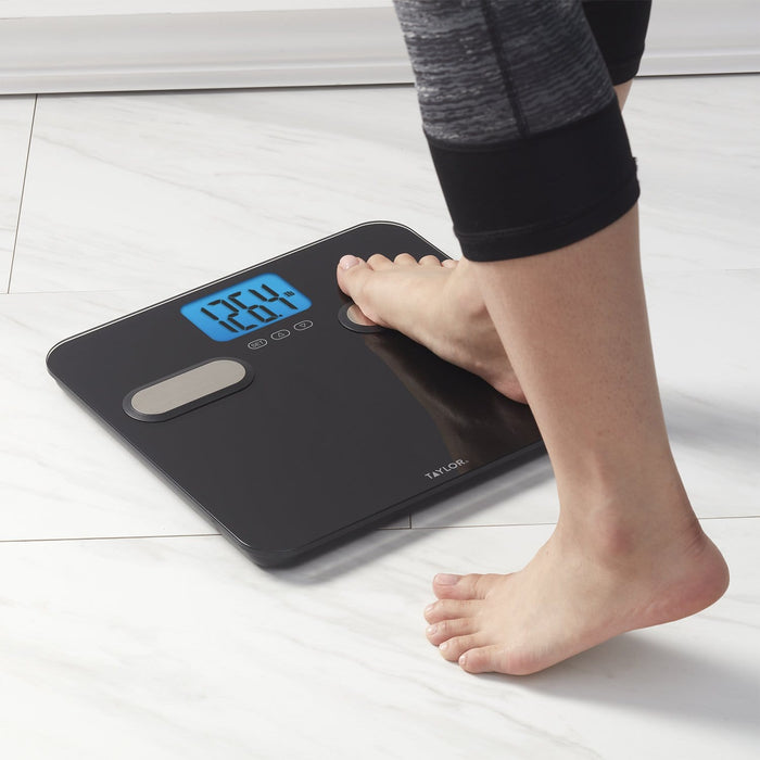 Person standing on a black Taylor body composition scale with blue backlit display on a light floor