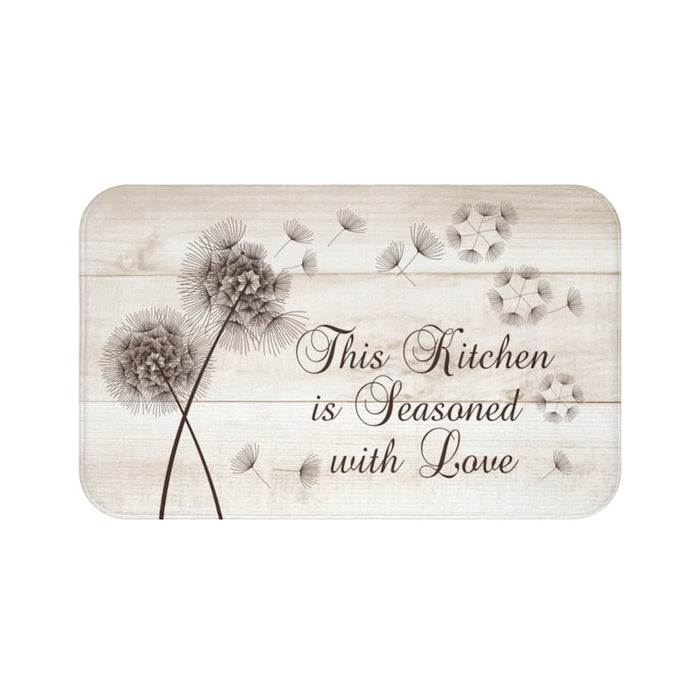 This is an image of a rectangular floor mat with rounded corners. The background of the mat mimics distressed, light-colored wooden planks. The horizontal plank lines give the mat a texture and farmhouse feel. The primary graphic is a cluster of stylized dandelion seed heads on the left side. The seed heads are drawn in fine detail, and several dandelion seeds are shown floating across the background, suggesting a gentle breeze with the phrase "This kitchen is seasoned with love".