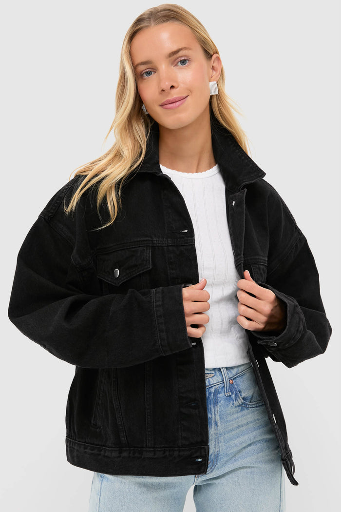 A woman with long blonde hair wears a Nude Lucy oversized vintage black stone wash denim jacket over a white ribbed sweater and light blue jeans.