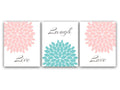 A set of three wall art prints with the words 'Live', 'Laugh', and 'Love' alongside corresponding pink and aqua floral burst designs. The left panel has a  soft pink floral burst filling the upper-right corner and the word "Live" in gray script at the bottom. The center panel has a the word "Laugh" in gray script at the top, and a large teal floral burst filling the bottom half. The right panel has a soft pink floral burst filling the upper-left corner and the word "Love" in gray script at the bottom.