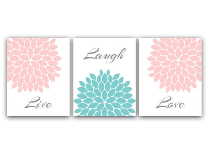 A set of three wall art prints with the words 'Live', 'Laugh', and 'Love' alongside corresponding pink and aqua floral burst designs. The left panel has a  soft pink floral burst filling the upper-right corner and the word "Live" in gray script at the bottom. The center panel has a the word "Laugh" in gray script at the top, and a large teal floral burst filling the bottom half. The right panel has a soft pink floral burst filling the upper-left corner and the word "Love" in gray script at the bottom.