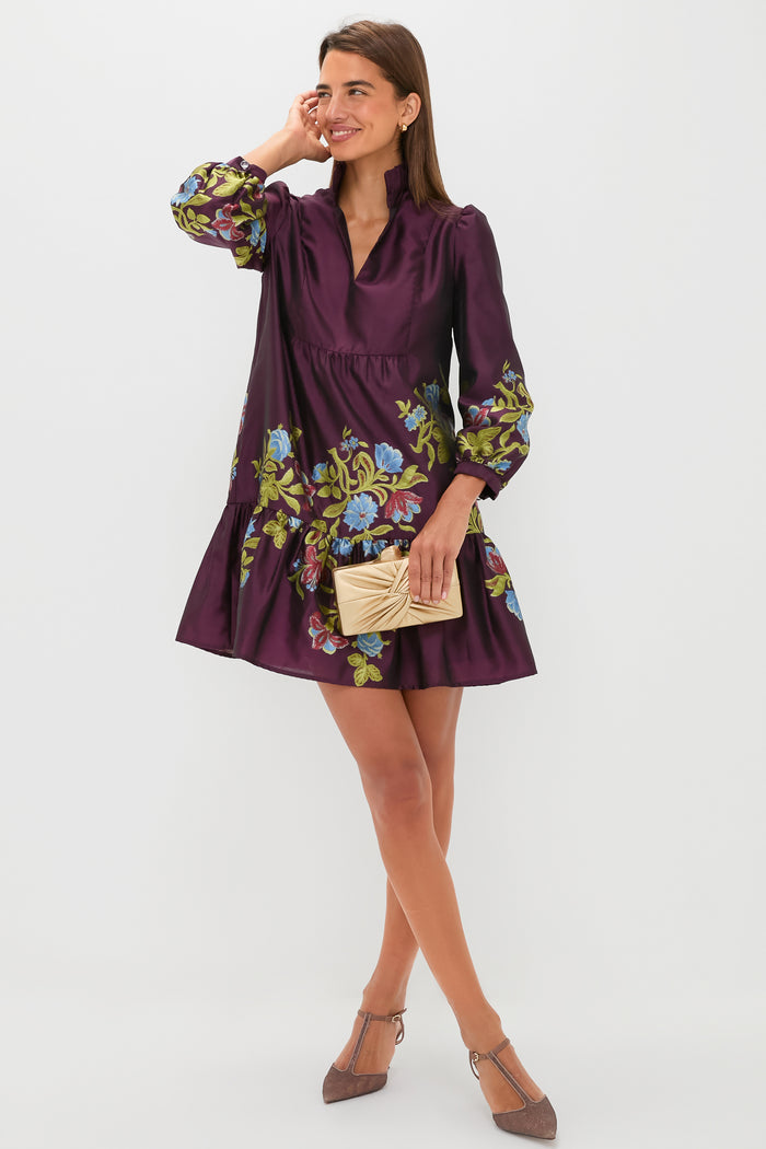 A woman wears the Tuckernuck Palmerston dress in Winetasting Dianthus Jacquard with a ruffle collar, holding a beige clutch and standing in neutral heels against a plain white background.
