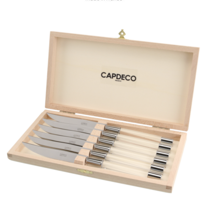 A wooden box with a hinged lid, branded Capdeco inside, is open to display six Zoe Steak Knives made in France, each with a steel blade and light-colored handle, all neatly arranged in individual slots.